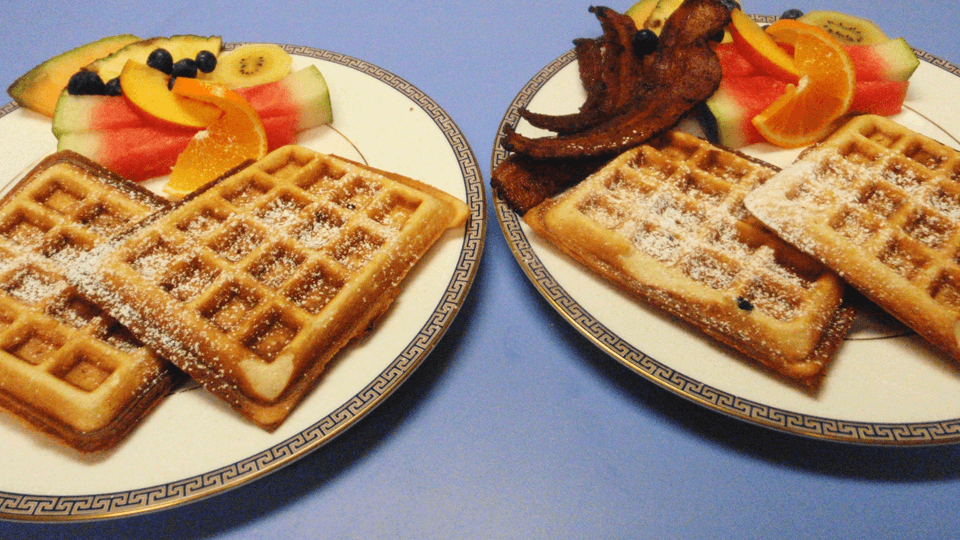 Two plates of waffles dusted with powdered sugar, accompanied by assorted fresh fruits and bacon.