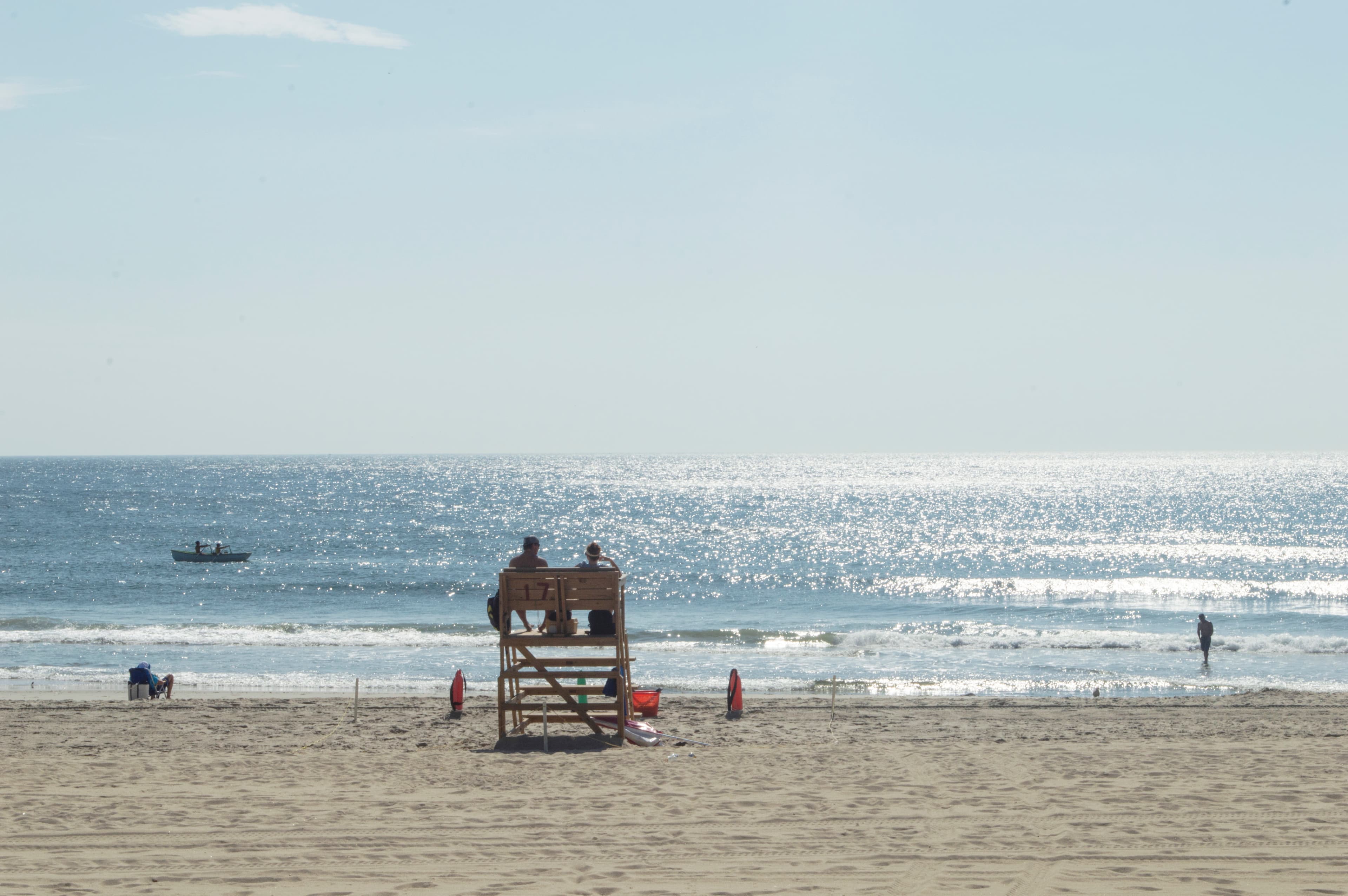 Two people sit on a lifeguard stand overlooking a sunny beach and glistening ocean.